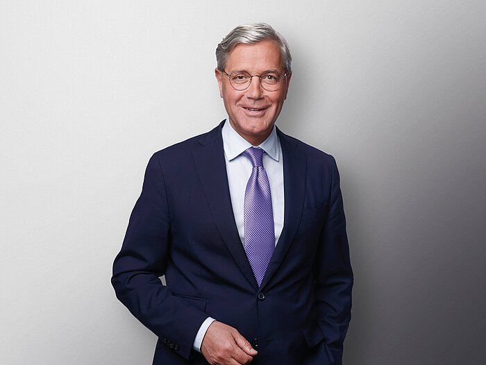 Das Bild zeigt Dr. Norbert Roettgen. Er lächelt freundlich und trägt ein dunkelblaues Jackett mit einer lila glänzenden Krawatte.
